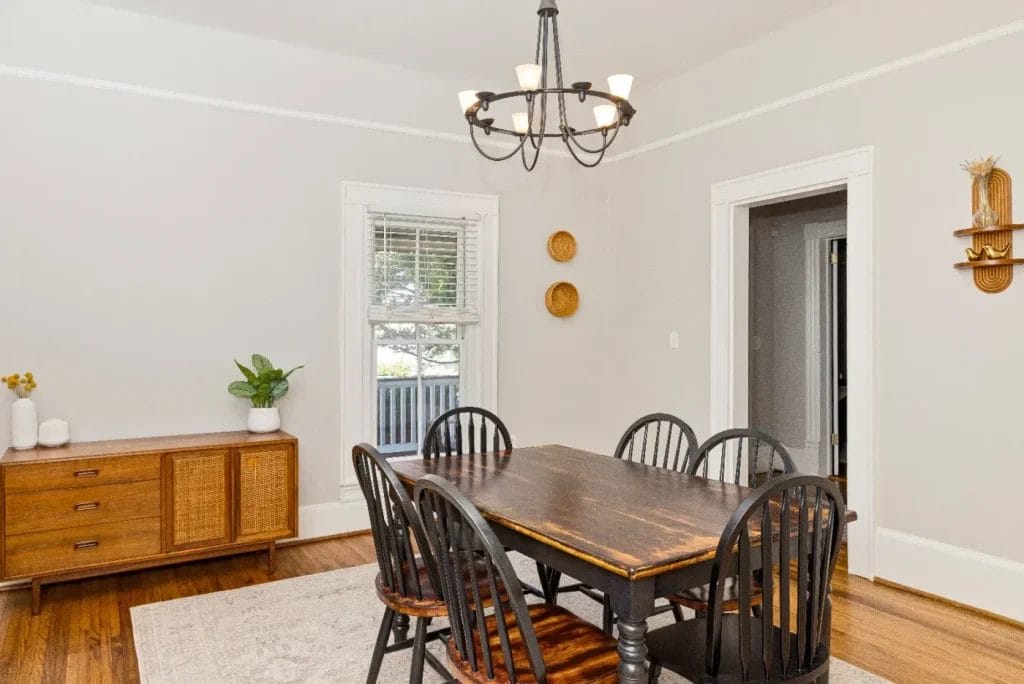 A bright dining room with a wooden table and six black chairs, a light fixture above, a wooden sideboard with plants and decor, and wall art accents on pale walls. A window lets in natural light.