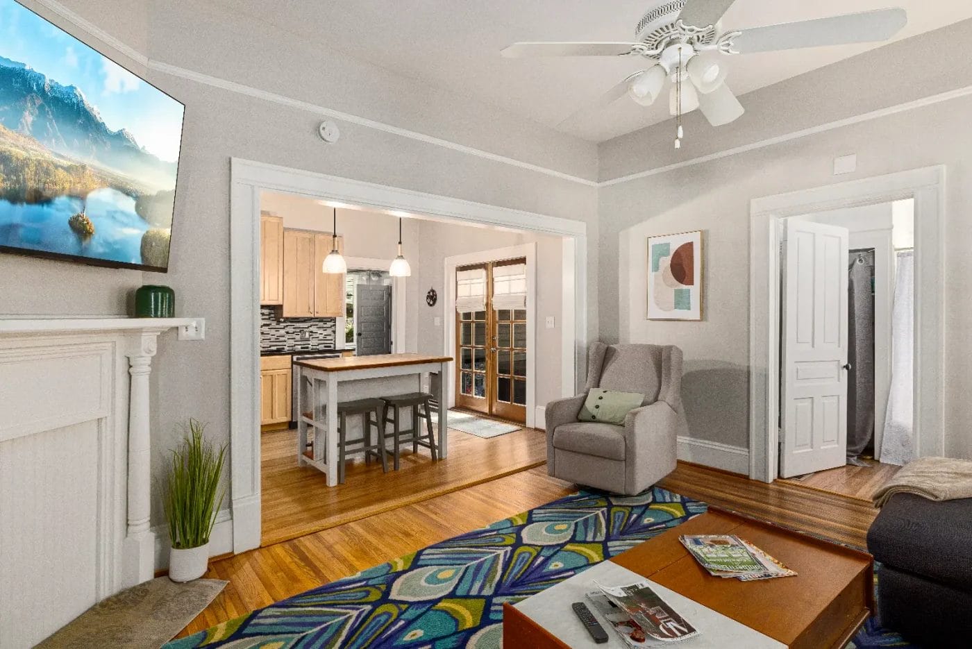 A cozy living room with a modern armchair, colorful rug, coffee table, TV mounted above a fireplace, and a view into a bright kitchen with a wooden island and stools. French doors let in natural light.
