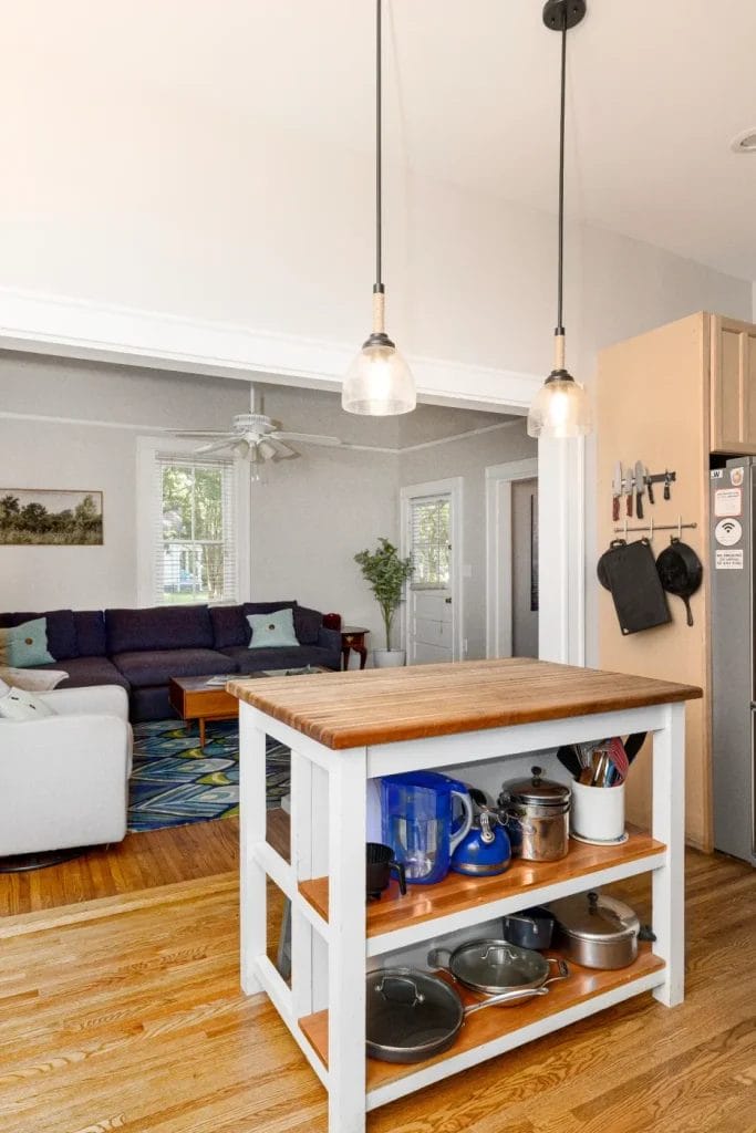 A bright kitchen features a wooden island with shelves holding pots, pans, and kitchenware. Pendant lights hang above, and a cozy living room with a sectional sofa is visible in the background.