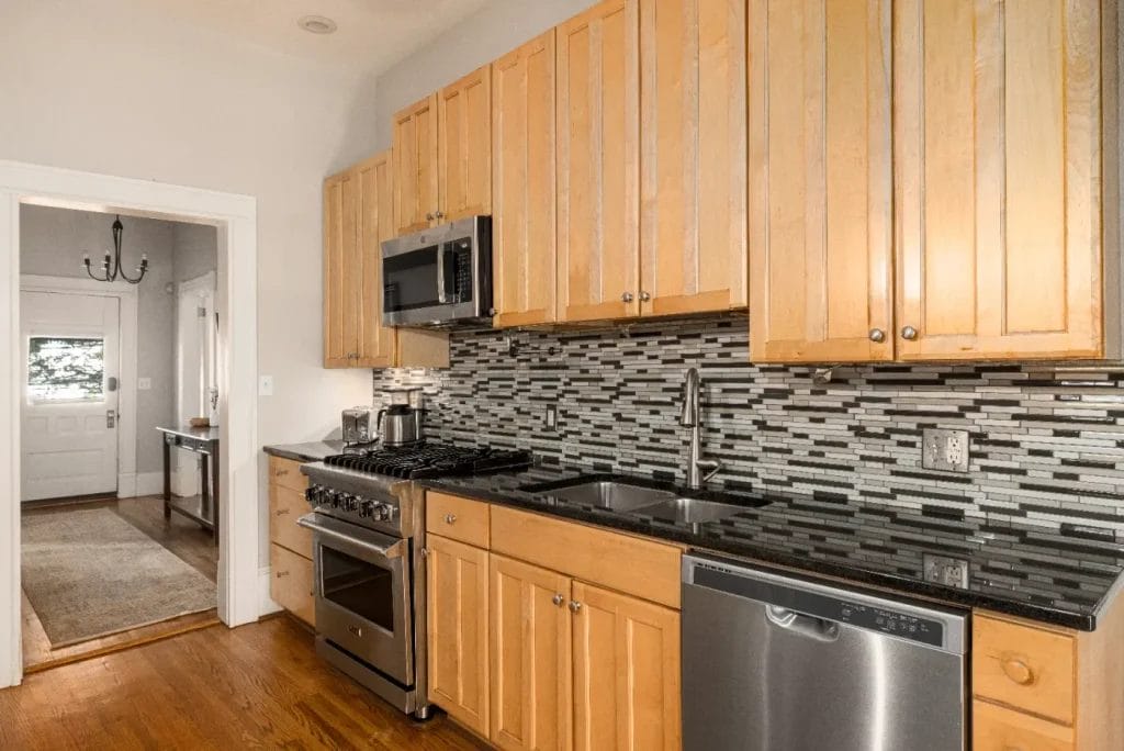 A modern kitchen with light wood cabinets, a black granite countertop, stainless steel appliances including a stove, microwave, and dishwasher, and a gray tile backsplash. A hallway and door are visible in the background.