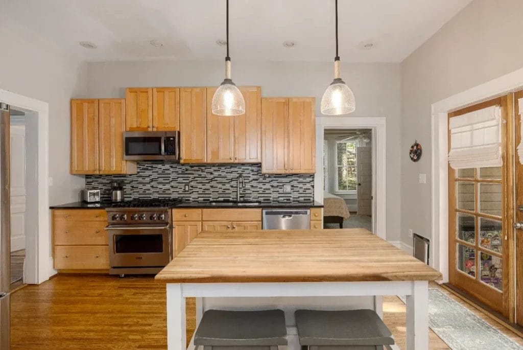 Modern kitchen with wood cabinets, stainless steel appliances, black countertops, tiled backsplash, and a wooden island with two gray stools. Pendant lights hang above the island, and double glass doors are on the right.