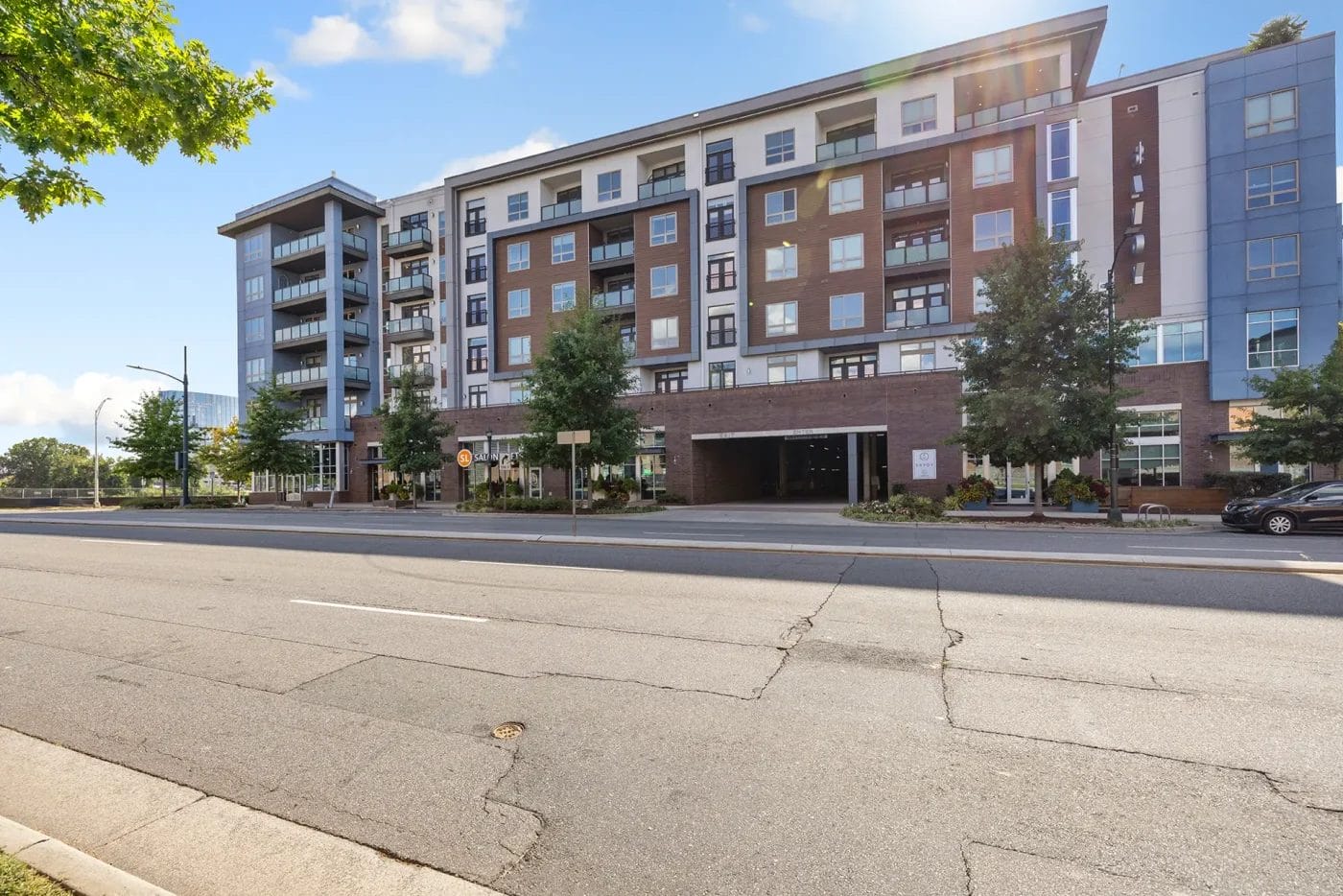 A modern multi-story apartment building with large windows, balconies, and a brick and siding exterior sits beside a quiet urban street with minimal traffic and trees along the sidewalk.