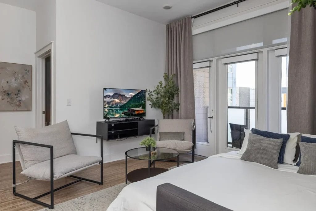 Modern bedroom with a bed, pillows, two cushioned chairs, a round glass coffee table, TV on a stand, potted plant, large windows with curtains, and light-colored walls and wood flooring.