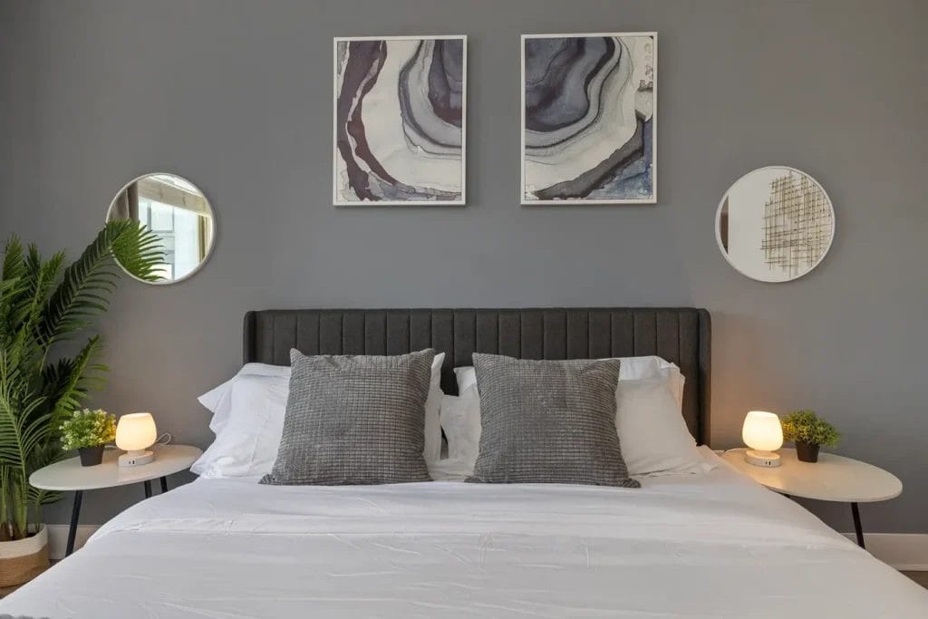 A modern bedroom with a gray upholstered headboard, white bedding, two gray pillows, and two round bedside tables holding lamps and small plants. Abstract art and round mirrors hang on a gray wall above the bed.