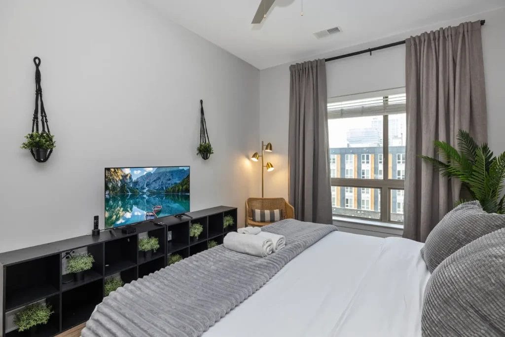 A modern bedroom with a neatly made bed, gray bedding, a TV on a black shelf with green plants, a wicker chair by the window, and gray curtains. Natural light fills the room.