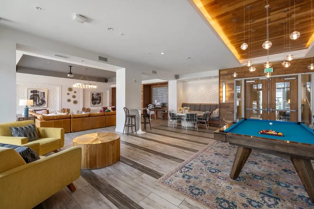 Modern lounge area with yellow and brown seating, a pool table, decorative lighting, wooden accents, and large windows providing natural light. The room has a relaxed, inviting atmosphere with contemporary decor.