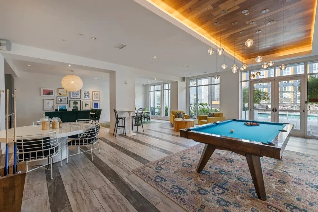 Modern lounge with a blue felt pool table, colorful rug, seating areas, wall art, and large windows overlooking a pool area outside. The room features wood and tile flooring and decorative pendant lights.