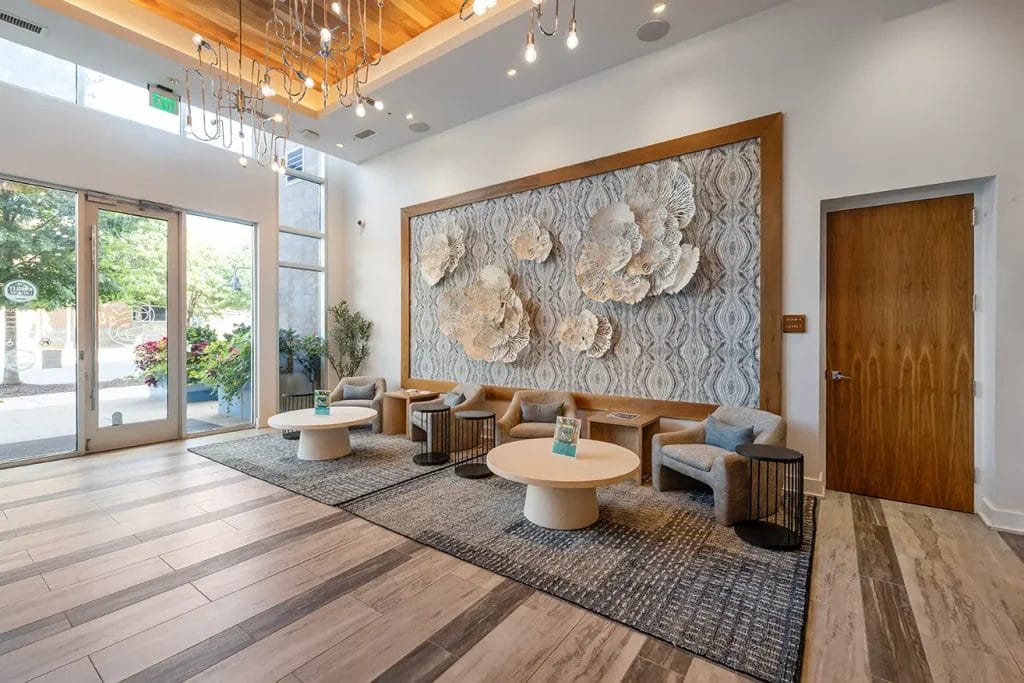 A modern lobby with wood flooring, five gray chairs around two light wood tables, large textured floral wall art, and a glass door leading outside to greenery. A chandelier hangs from the wooden ceiling accent.