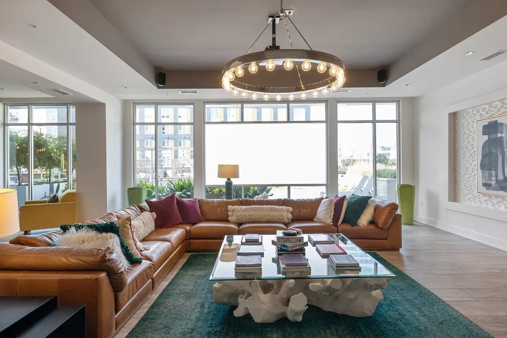 Modern living room with a large brown leather sectional sofa, colorful pillows, glass coffee table, teal rug, and a circular chandelier. Floor-to-ceiling windows provide natural light and a cityscape view.