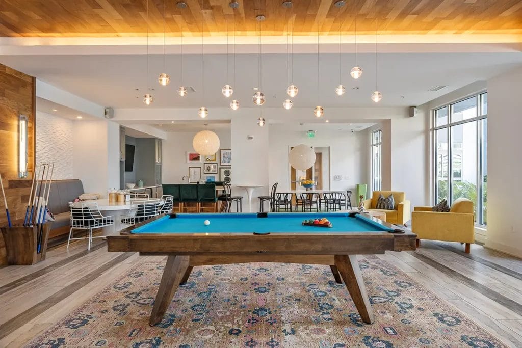 A modern lounge with a blue pool table centered on a patterned rug, surrounded by stylish seating, dining tables, and large windows letting in natural light. Spherical pendant lights hang from a wooden ceiling feature.