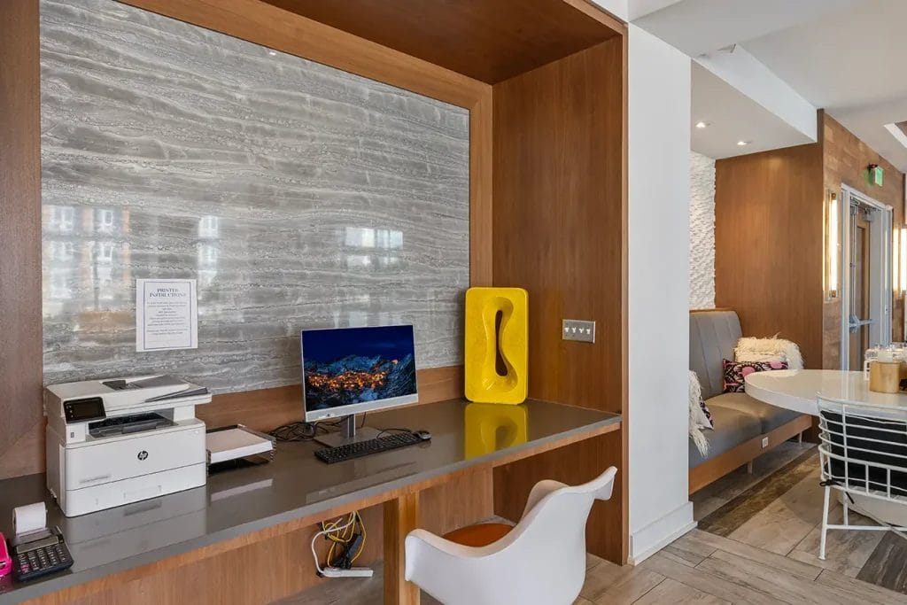A modern workspace with a computer, keyboard, mouse, and printer on a sleek desk. A yellow decorative piece and a white chair are next to the setup, with wood paneling and gray stone accent wall behind.