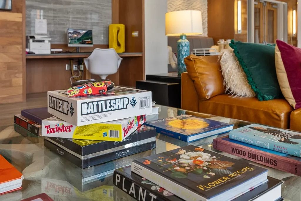 A glass coffee table holds board games like Battleship and Twister, and books about plants, flowers, and wood buildings. In the background is a cozy seating area with a desk, chair, and a tan couch with colorful pillows.