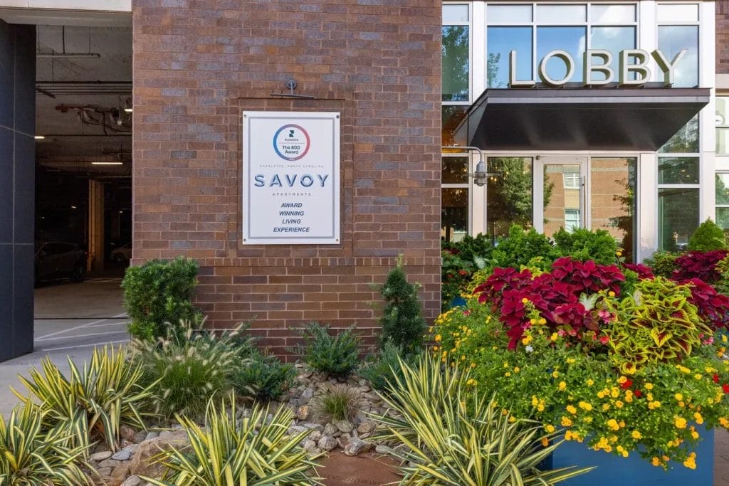 A brick building entrance with a "LOBBY" sign, lush landscaping, colorful flowers, and a plaque reading "Savoy Apartments Award Winning Living Experience.