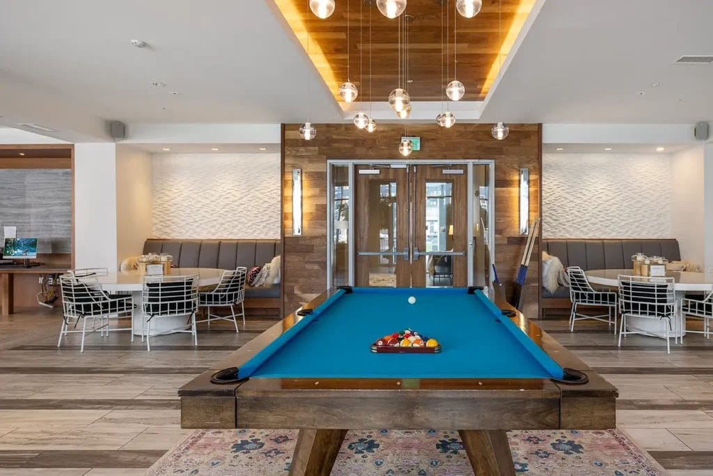 A modern game room with a blue pool table at the center, stylish pendant lights above, wood accents, and seating areas with tables and chairs along the walls.