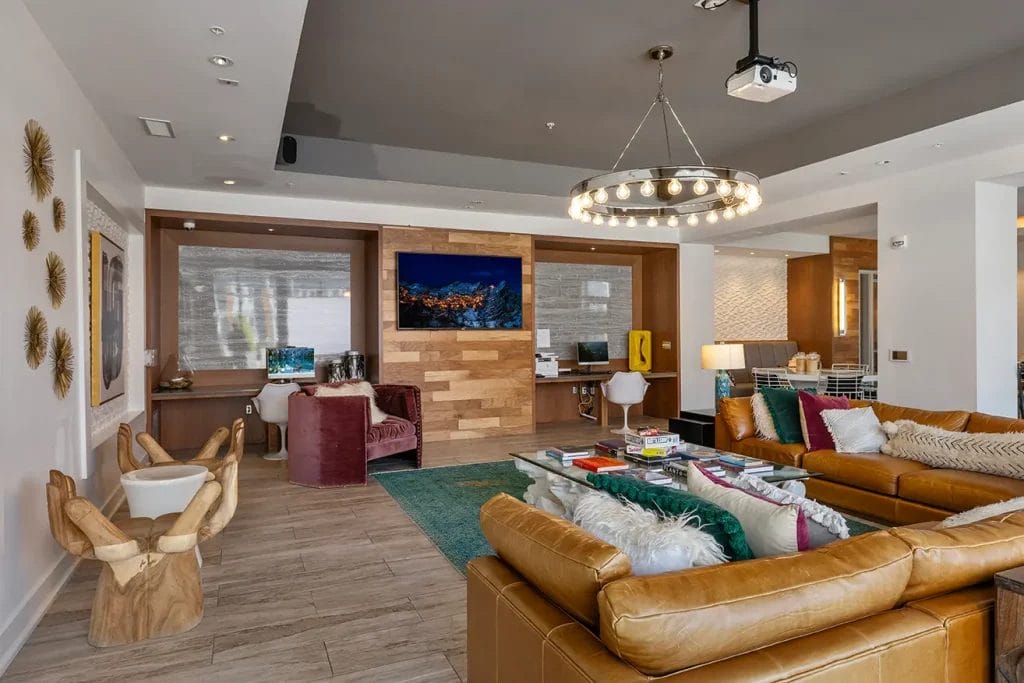 A modern living room with tan leather sofas, colorful pillows, a wood accent wall with a TV, a circular chandelier, a green rug, office desks with computers, and various decor elements, including wall art and a sculptural chair.