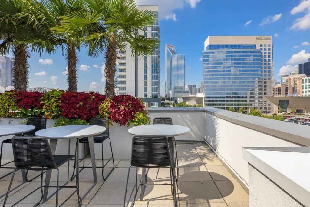 Outdoor rooftop patio with black chairs and round white tables, lined with palm trees and red-leaved plants. Tall modern buildings and a clear blue sky are visible in the background.