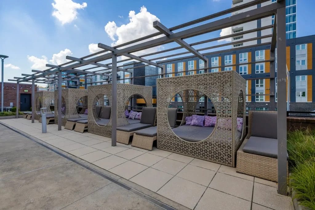 Outdoor lounge area with stylish cabanas featuring circular openings, modern wicker furniture, and purple-patterned pillows. Colorful apartment buildings and a blue sky with clouds are in the background.