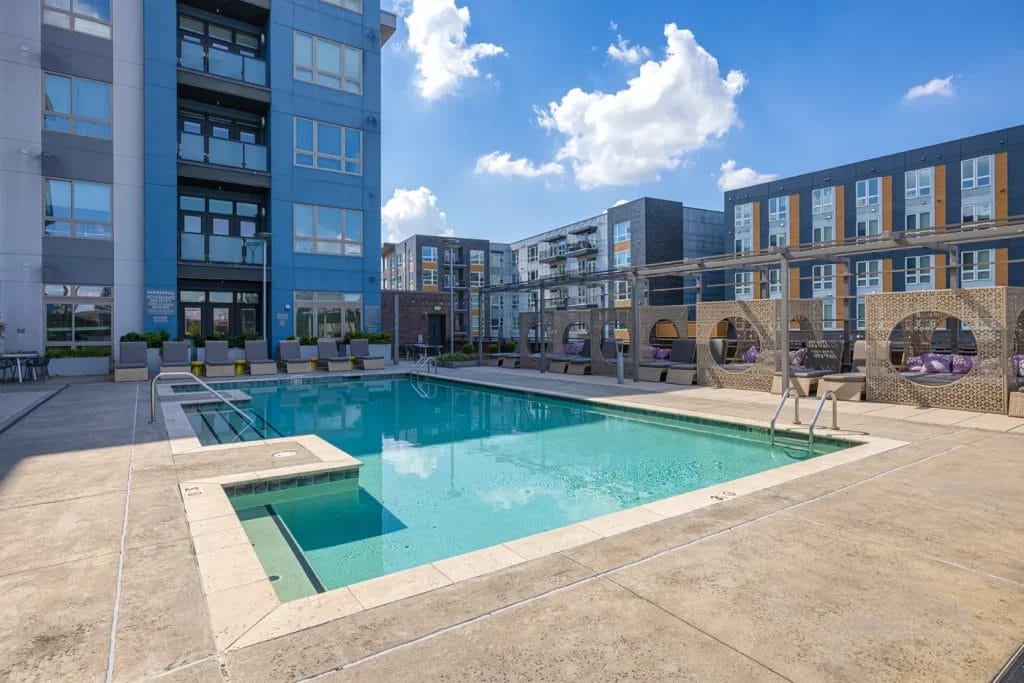 A modern apartment complex features a clean outdoor swimming pool with clear blue water, surrounded by lounge chairs and shaded cabanas under a sunny sky with a few clouds.