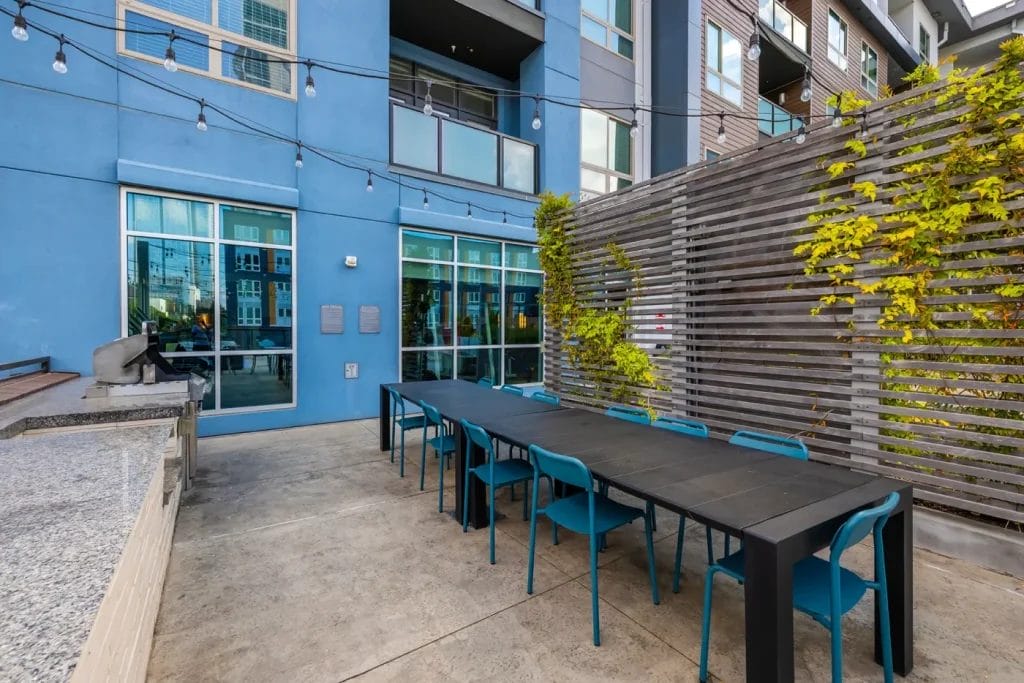 Outdoor patio area at a modern apartment complex with a long black table, blue chairs, string lights, and a wooden privacy screen with climbing plants. Large windows and blue building facade in the background.