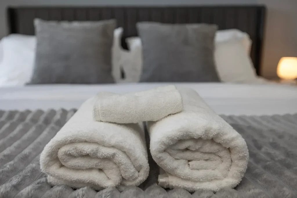 Two neatly rolled white towels with a folded washcloth on top are placed on a bed with a gray textured blanket. In the background, there are several gray and white pillows arranged against the headboard.