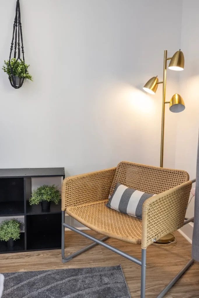 A cozy corner with a wicker chair and striped pillow, next to a gold floor lamp with three lights. A black cubby shelf with potted plants and a hanging plant decorate the space.