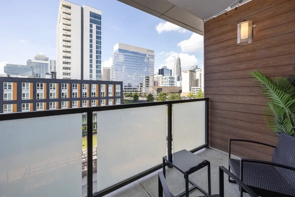 A modern balcony with frosted glass railing, black chairs, a small table, a potted plant, and a city skyline with high-rise buildings in the background under a blue sky.