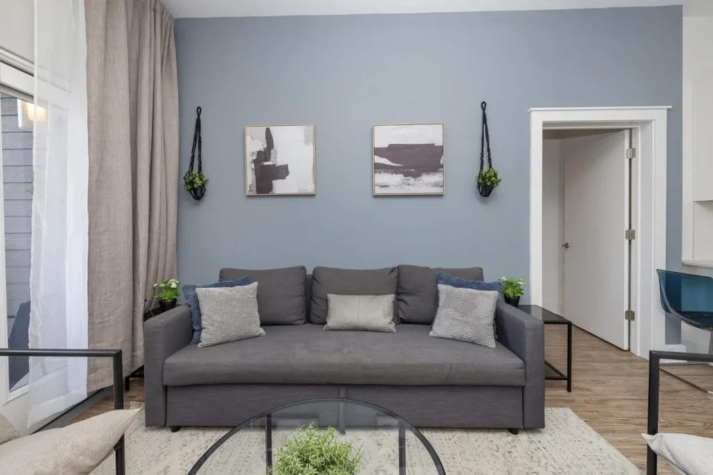 A modern living room with a gray sofa, three pillows, two abstract paintings on a blue-gray wall, hanging plants, a glass coffee table, neutral curtains, and a partially open door to the right.