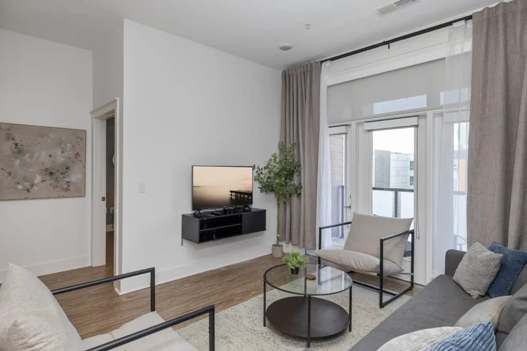 Modern living room with a wall-mounted TV, two armchairs, a gray sofa, a round glass coffee table, light wood floors, sheer curtains, and large windows leading to a balcony.