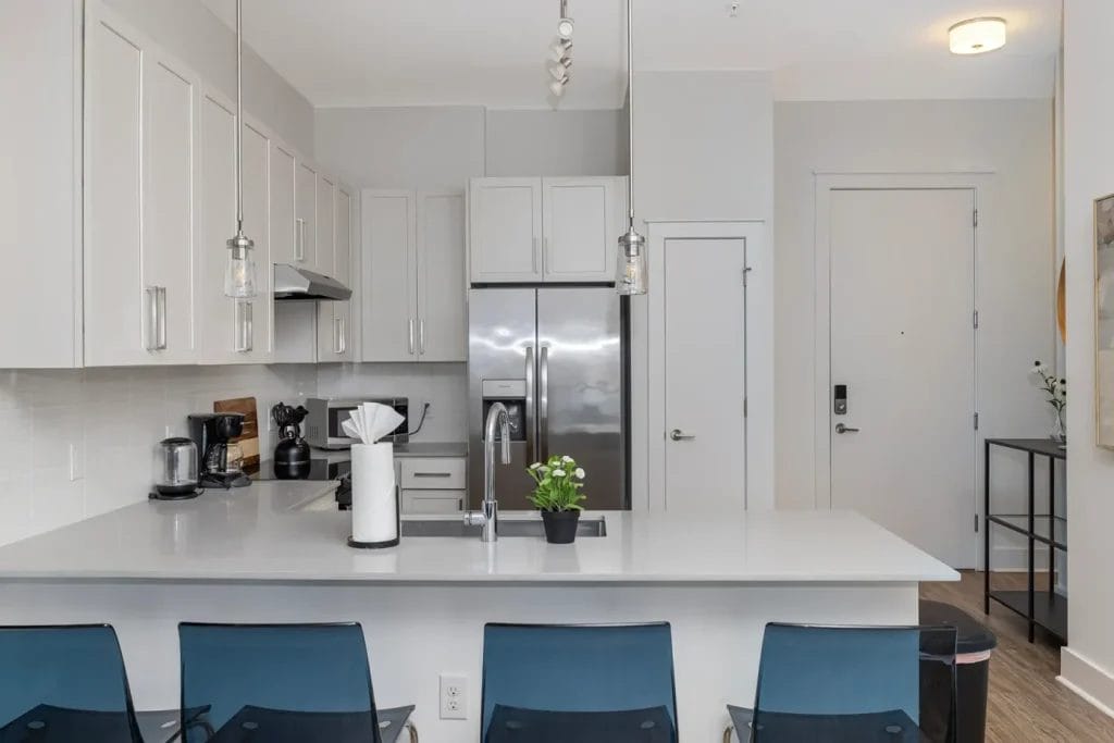 Modern kitchen with white cabinets, stainless steel appliances, a large island with a small potted plant, blue chairs, pendant lights, and a paper towel roll on the counter.