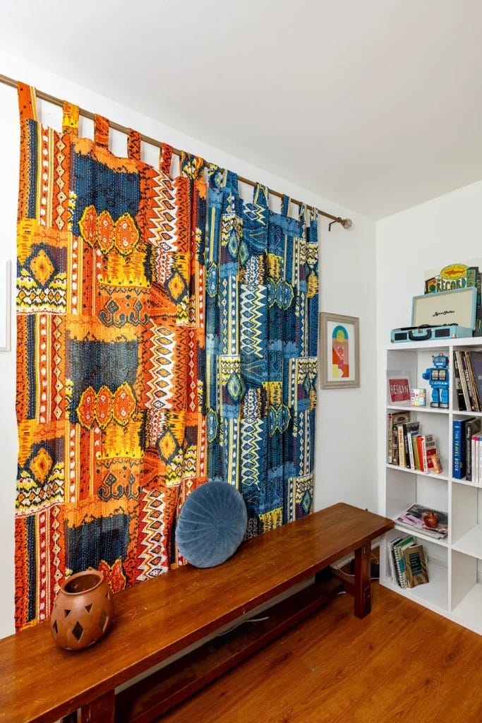 A colorful airbnb room features vibrant patterned curtains in orange and blue, a wooden bench with a round blue cushion, a lantern, and framed art—perfectly captured by an Artistic Photographer in Charlotte NC for stylish stays.