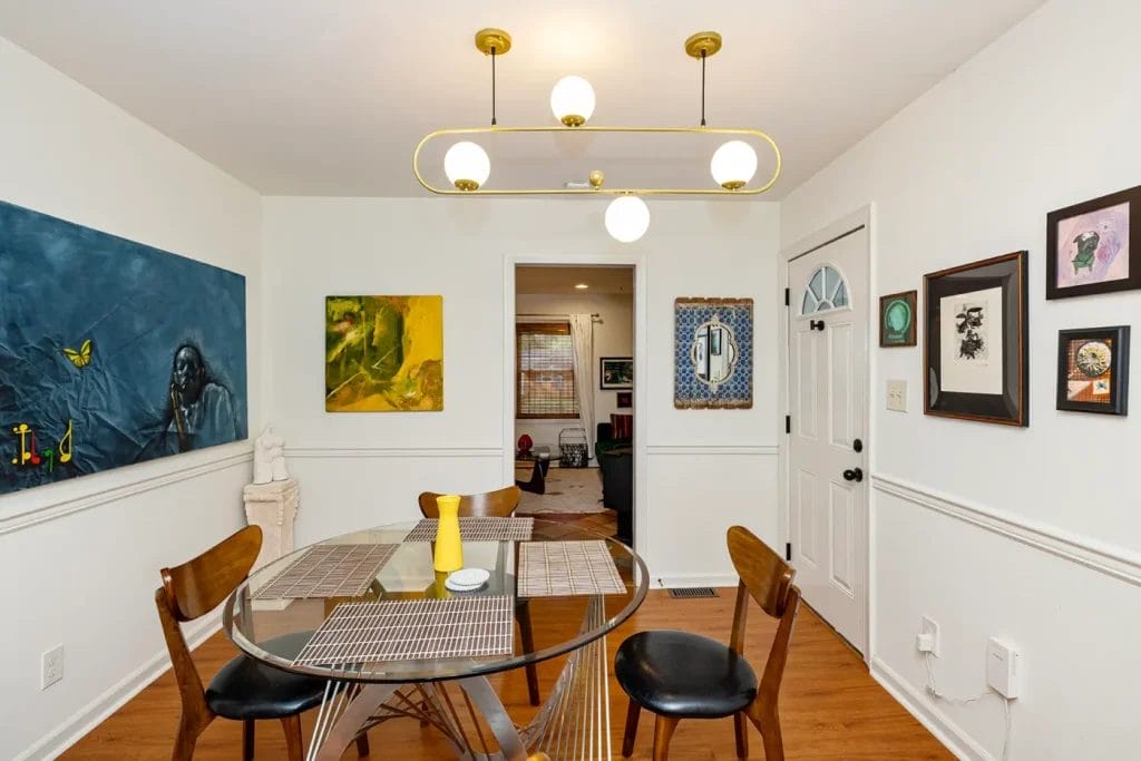 A modern dining room with a glass table, four wooden chairs, contemporary light fixture, and various artworks on the white walls—captured by an Artistic Photographer in Charlotte NC. A yellow vase sits on the table, and two doorways lead to other rooms.
