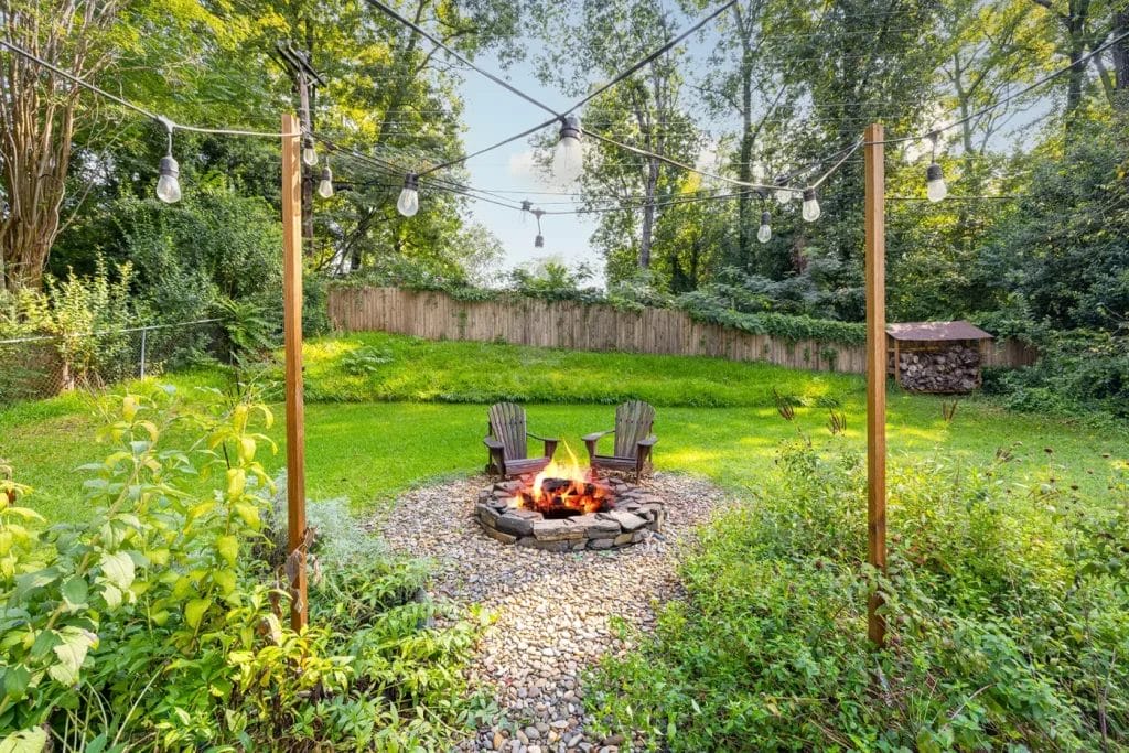A backyard with a stone fire pit surrounded by two wooden Adirondack chairs, string lights overhead, lush green grass, trees, and a wooden fence—beautifully captured by an artistic photographer in Charlotte NC for Airbnb listings.