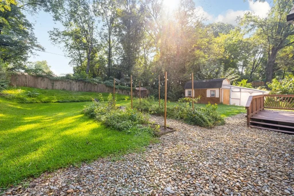 Sunny backyard with green grass, a small garden with stakes, wooden deck, gravel area, wooden fence, and two sheds surrounded by tall trees—perfectly captured by an Artistic Photographer in Charlotte NC for your next Airbnb listing.