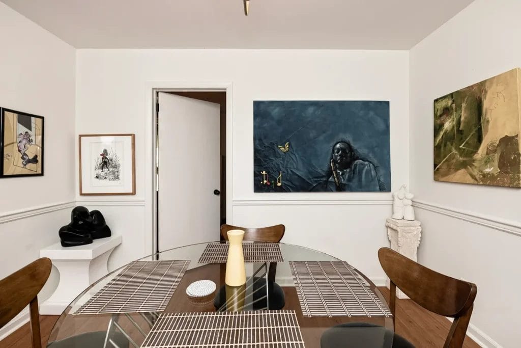 A modern dining room with a glass table, four wooden chairs, placemats, and a yellow vase. Contemporary art adorns white walls and sculptures sit on pedestals—perfectly staged for an airbnb photographer seeking artistic shots.