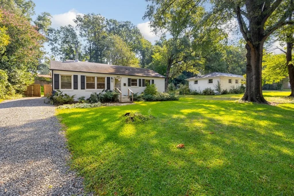 A single-story white house with a front porch and large windows sits on a green lawn with trees and shrubs. A gravel driveway runs along the left side, and a detached white garage is visible in the background.