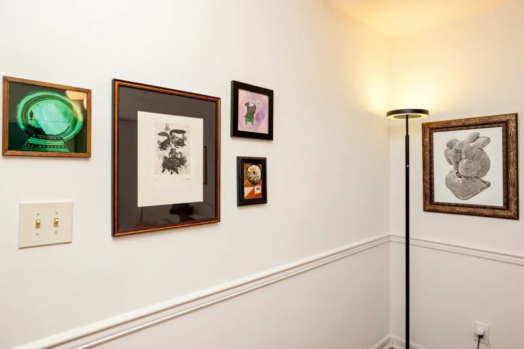 A corner of a room with five framed artworks on white walls, a black floor lamp with a round shade, a light switch, and white trim. Captured by an Artistic Photographer in Charlotte NC, the art features abstract and illustrative designs.