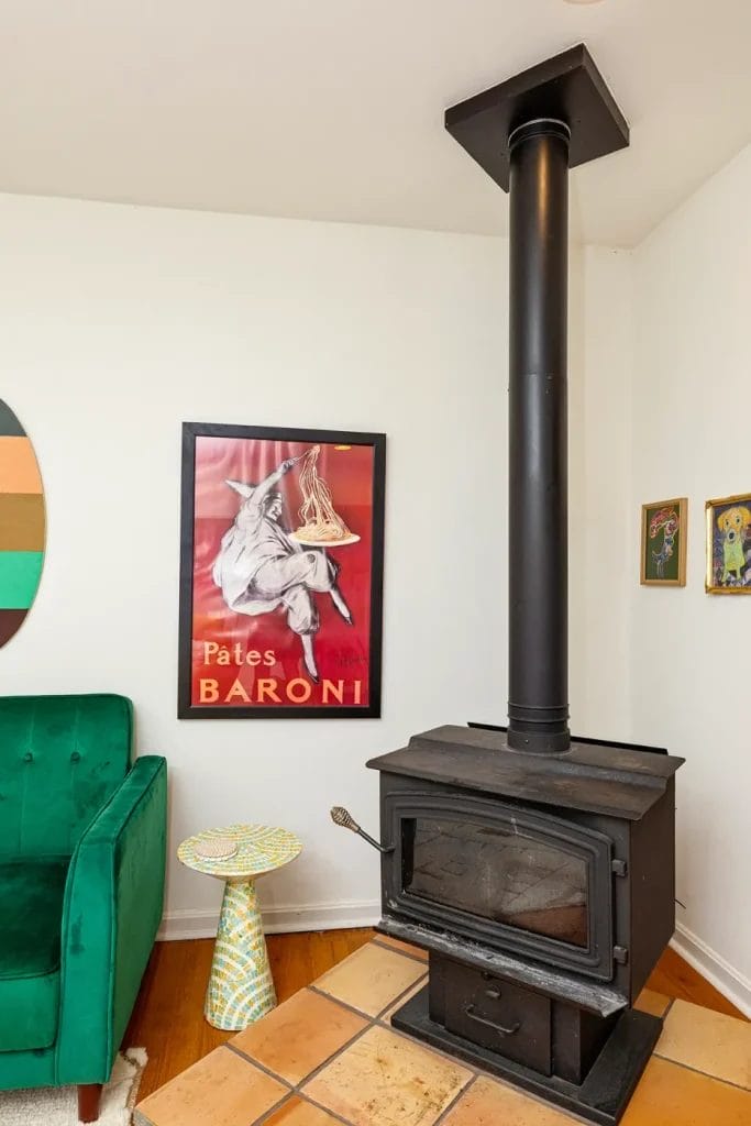 A black wood-burning stove with a tall chimney stands on a tiled floor next to a green velvet chair, a mosaic side table, and a framed vintage "Pâtes Baroni" poster—captured by an artistic photographer in Charlotte NC for your airbnb inspiration.