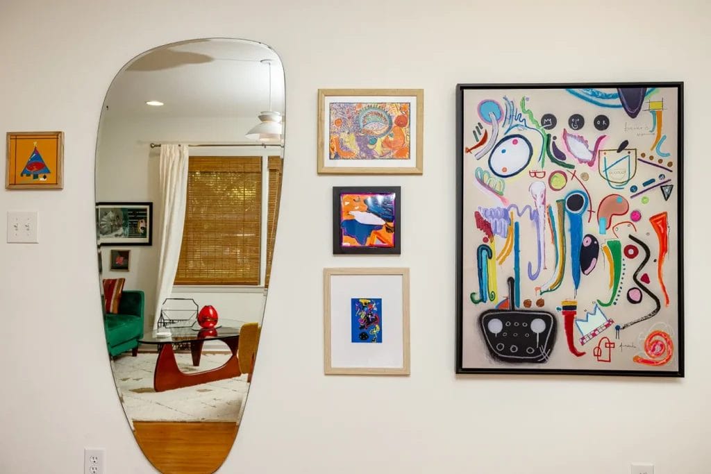 Artistic Photographer in Charlotte NC 23 A wall displays colorful abstract art and small framed prints, curated by an Artistic Photographer in Charlotte NC, next to an irregular mirror reflecting a living room with a green couch, red lamp, blinds, and curtain—ideal for an Airbnb listing.