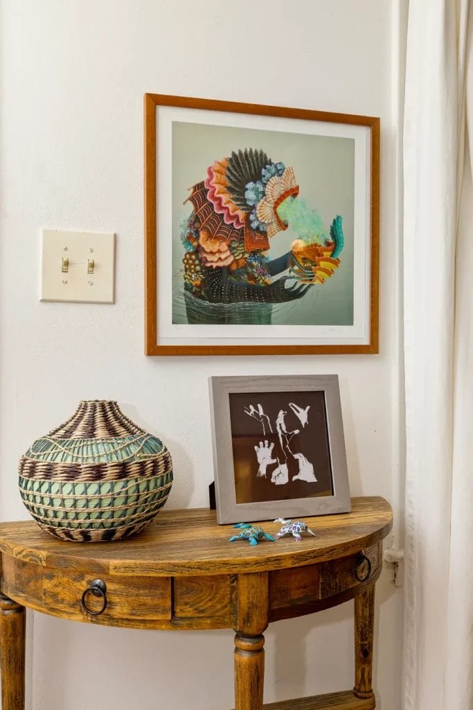 A wooden table with a woven vase, two small animal figurines, and a framed silhouette artwork sits below a colorful fish picture on a white wall near a window—perfect for an Airbnb listing by an artistic photographer in Charlotte NC.