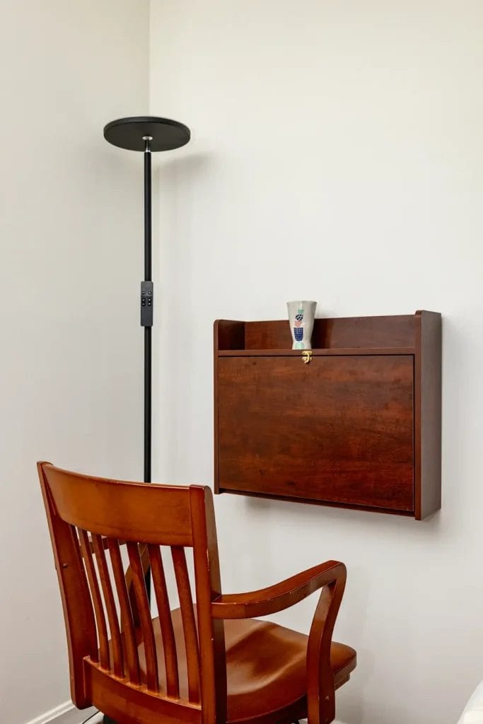 A wooden chair sits before a wall-mounted wooden desk, with a disposable cup on top—perfectly styled for an airbnb listing. A tall black floor lamp stands to the left, all set against a plain white wall.