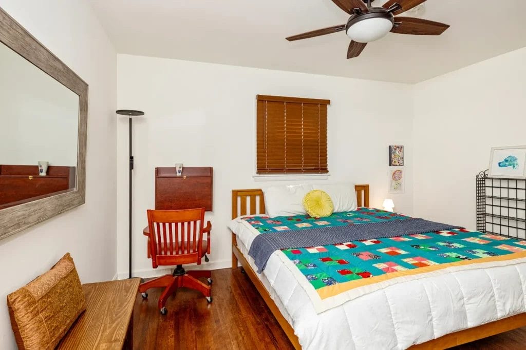 A cozy bedroom, perfect for Airbnb, with a wooden bed, colorful quilt, yellow round pillow, ceiling fan, red office chair, floor lamp, wall art, large mirror, and a small bench—captured by an artistic photographer in Charlotte NC.