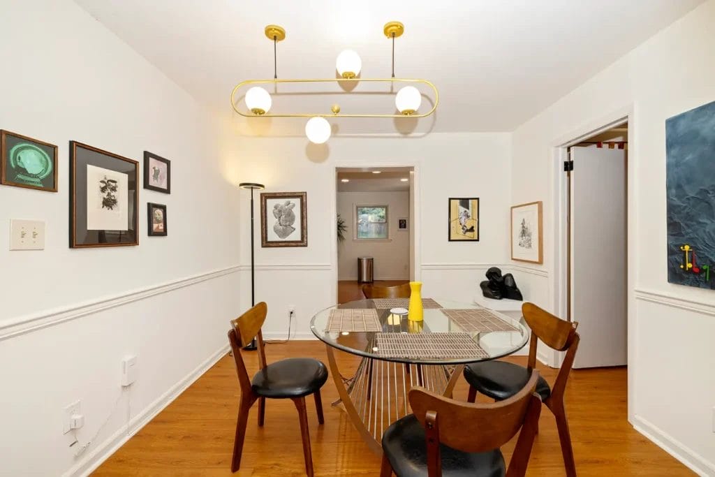 A modern dining room, ideal for an Airbnb listing, features a round glass table, four wooden chairs, a yellow vase, framed art on white walls, and a gold chandelier. Captured by an Artistic Photographer in Charlotte NC through an open doorway.