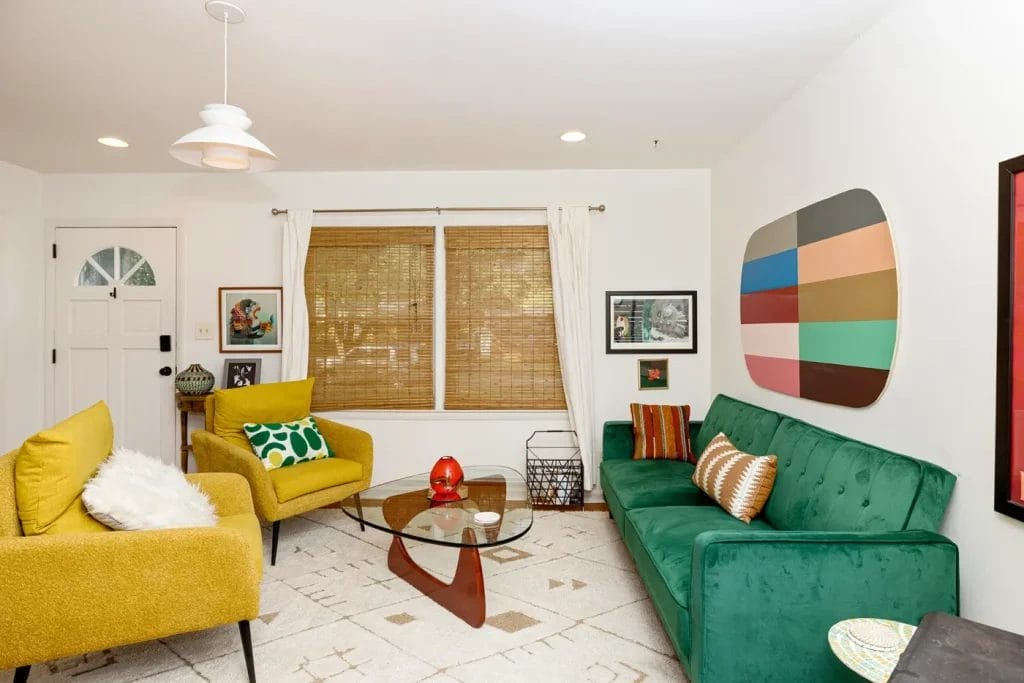 A modern living room perfect for Airbnb, featuring a green velvet sofa, two yellow armchairs, geometric wall art, and large windows with bamboo shades—ideal for an artistic photographer in Charlotte NC to capture vibrant, mid-century style.
