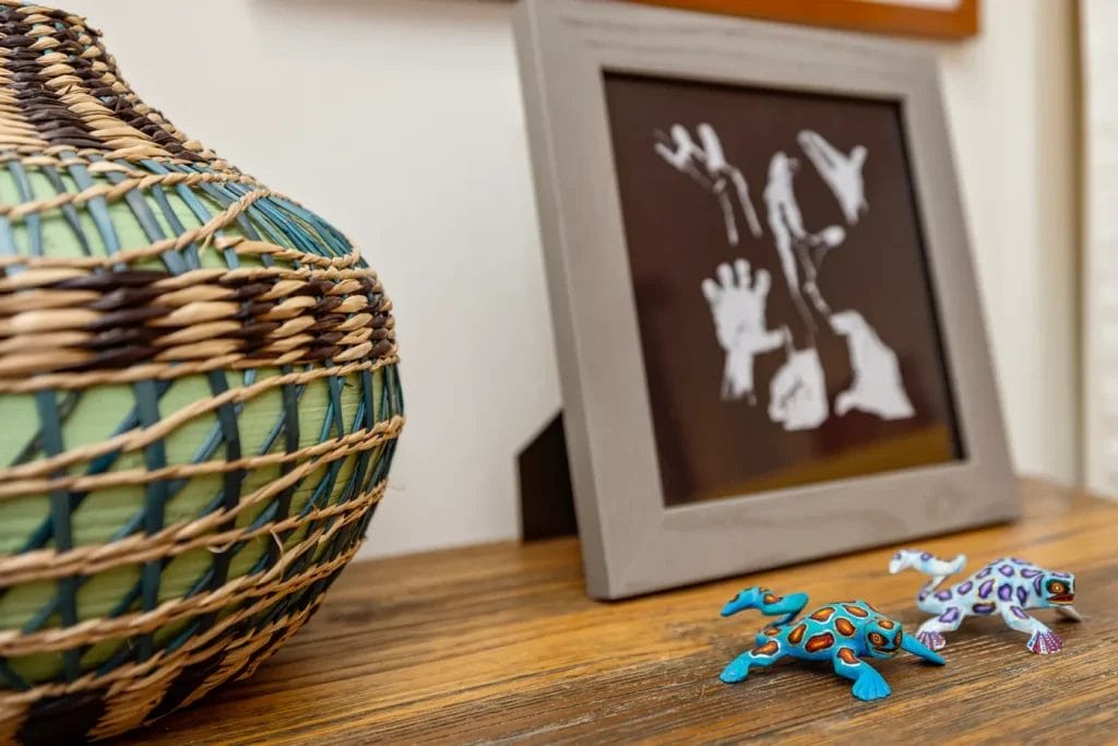 A woven basket, a framed art piece with white handprints, and two colorful ceramic frog figurines are artfully arranged on a wooden surface by an artistic photographer in Charlotte NC.