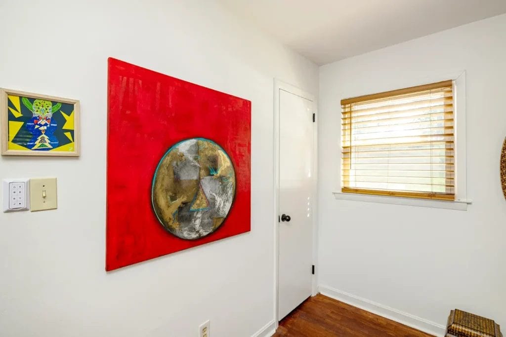 A small, white room with wooden floor, a window with bamboo blinds, a closed white door, and two colorful abstract art pieces—captured by an Artistic Photographer in Charlotte NC—perfect for an inspiring Airbnb stay.
