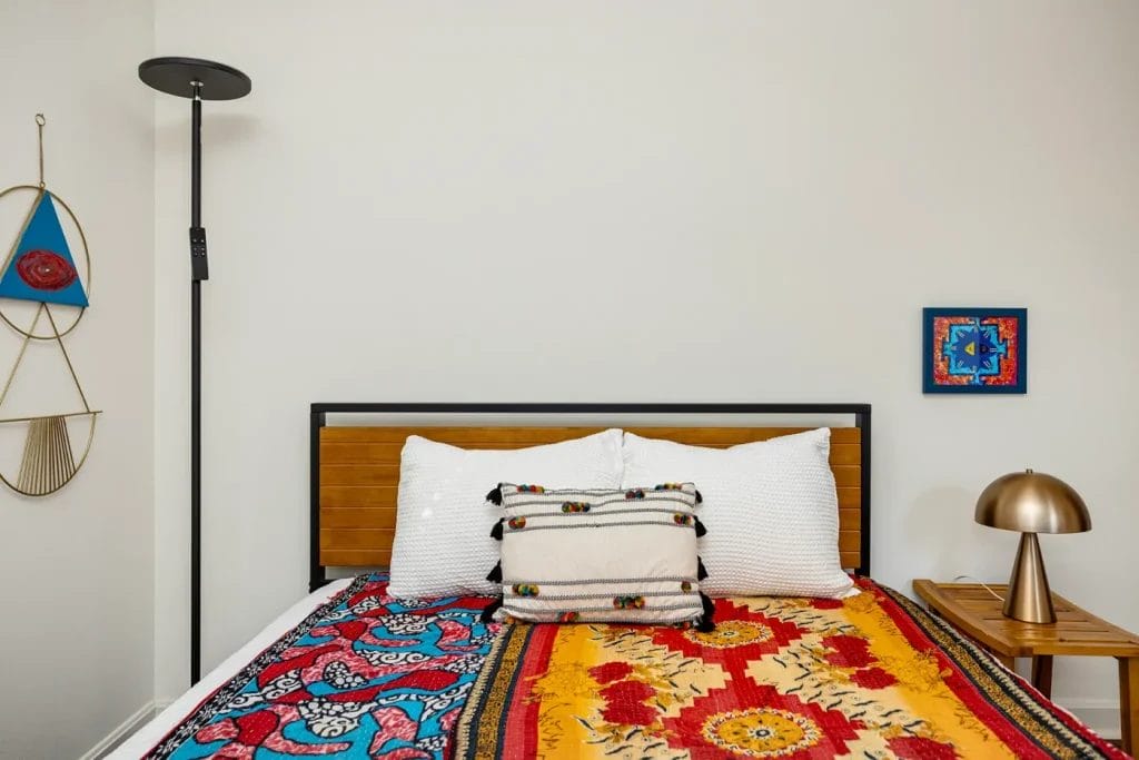 A neatly made bed with colorful, patterned blankets, white pillows, and a decorative cushion. Captured by an Airbnb photographer, the scene features modern lamps, a wooden nightstand, and abstract wall art for a welcoming touch.