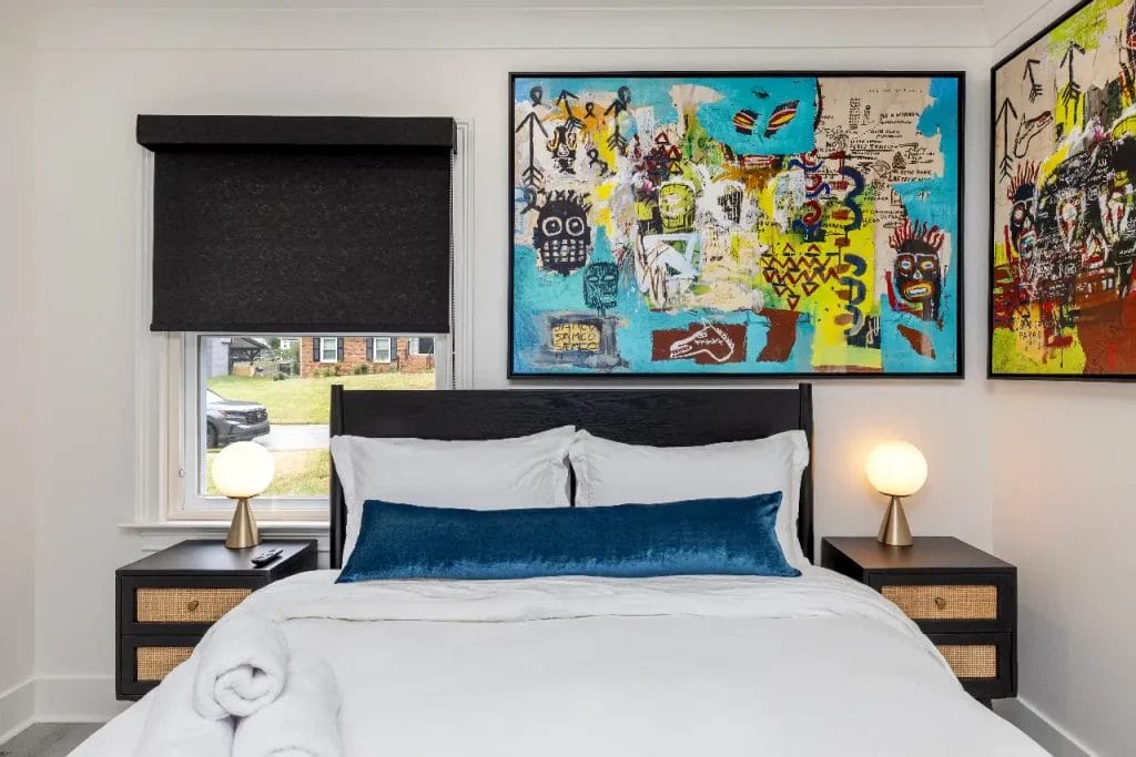 A neatly made bed with white linens and a blue accent pillow sits between two black nightstands with globe lamps. A large, colorful abstract painting hangs above the bed. A window with a black shade is on the left.