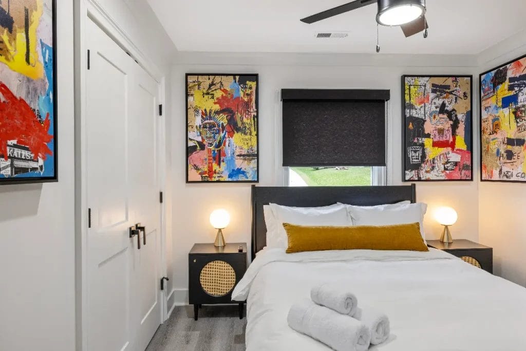 A modern bedroom with a double bed, white bedding, a yellow accent pillow, and two rolled towels. Nightstands with round lamps flank the bed, and colorful abstract art hangs on the walls. A window with a black shade is above the bed.