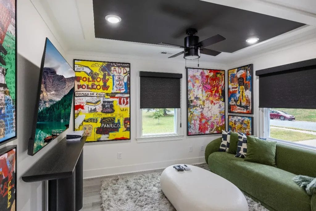 A modern living room with colorful, abstract art on the walls, a green couch with patterned pillows, a white oval coffee table, a textured rug, and a wall-mounted TV. Large windows let in natural light.