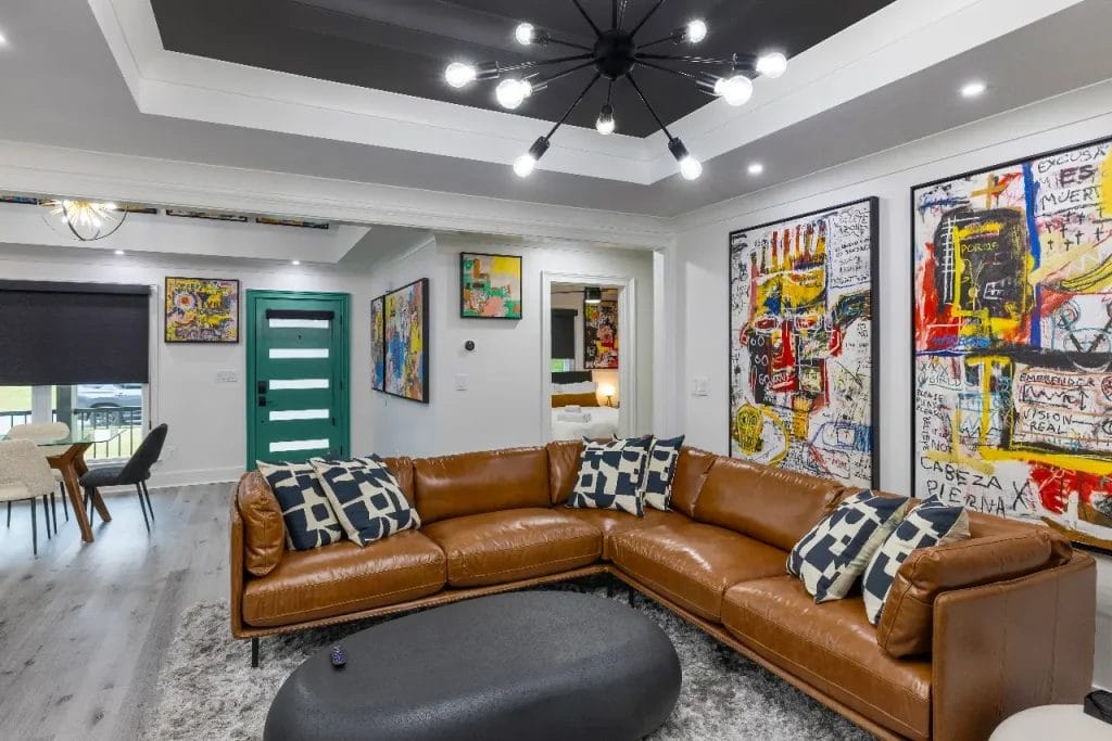 Modern living room with a large brown leather sectional sofa, geometric patterned pillows, abstract colorful wall art, a black oval coffee table, and a bold black chandelier with multiple bulbs.